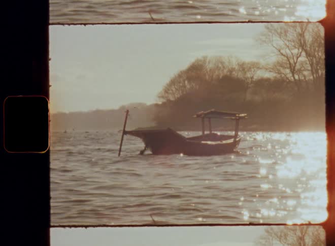 The empty boat is floating in the water moving in the ocean waves with the reflection of bright sunshine blinding and glaring on the rippling water surface by the coast or shore with trees in the background