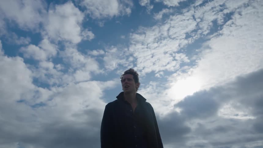Man kicks at sand while looking lost and upset on ground near water across from mountains on cloudy day