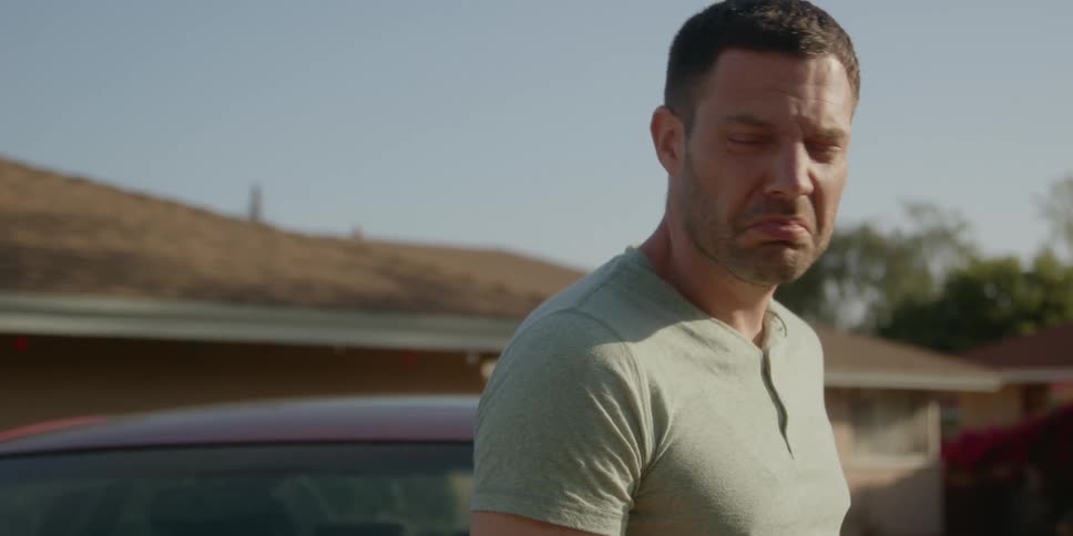 A man standing next to his car in front of a house engaged in conversation with someone while smoking a cigarette