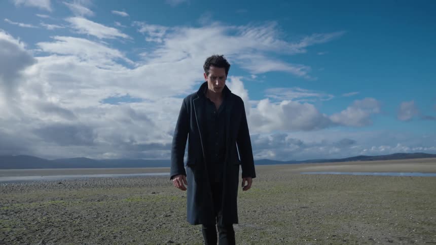 A man in a long coat walks through a vast field during the day