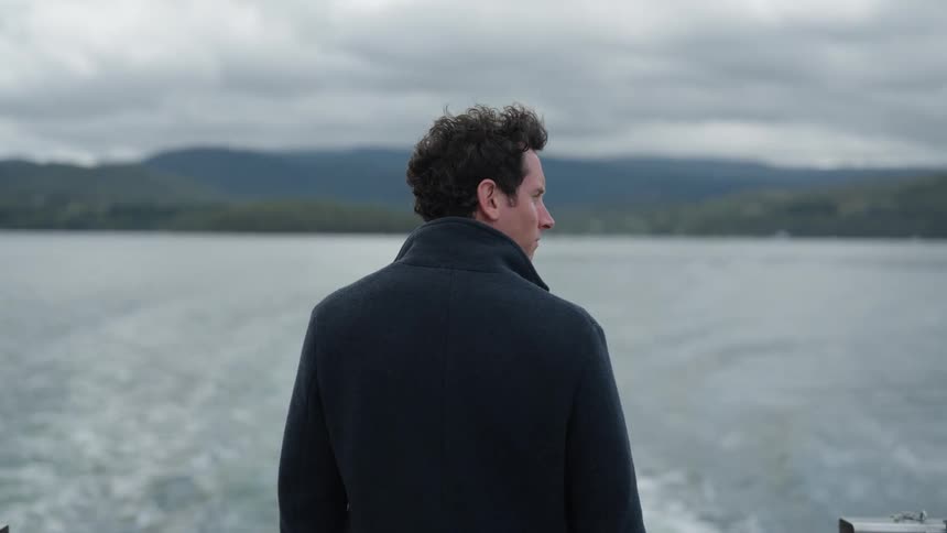 Man stands in back of motorboat and watches retreating coast on overcast day