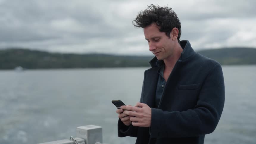 Man standing in back of boat speeding away from shore smiles and texts