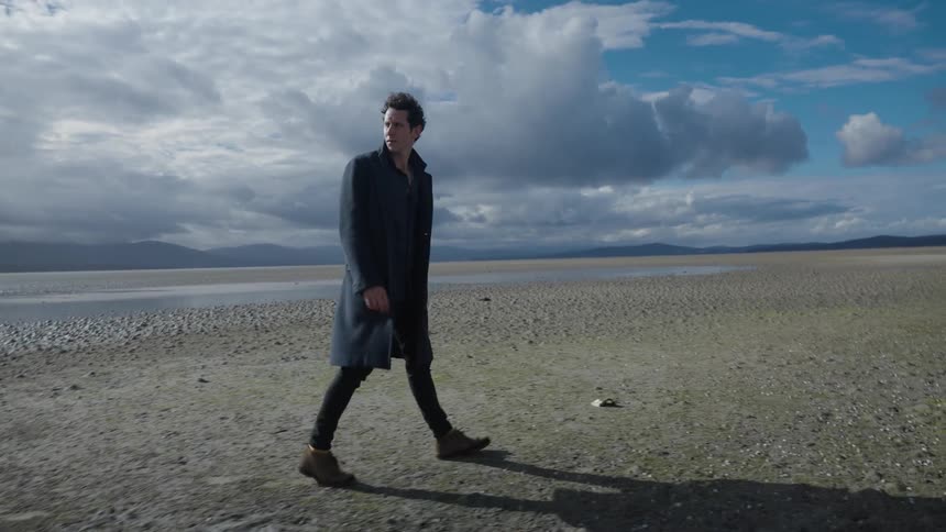 A man wearing an overcoat walking alone through a desertlike open field during the daytime