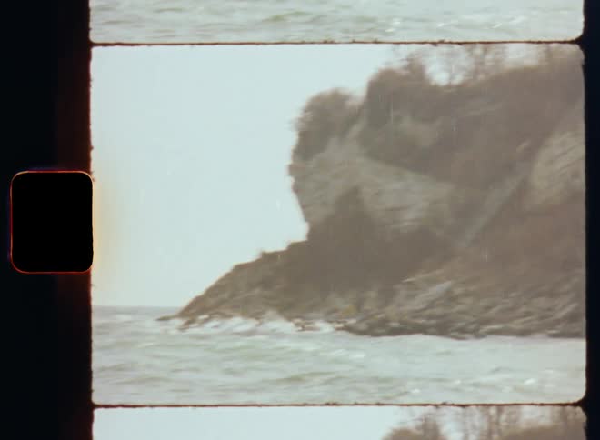 Film footage of a house sitting above a cliff on the shore as wave roll in