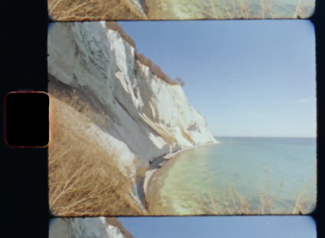 Narrow strip of beach separates white cliffs dotted with yellow vegetation from pale sea