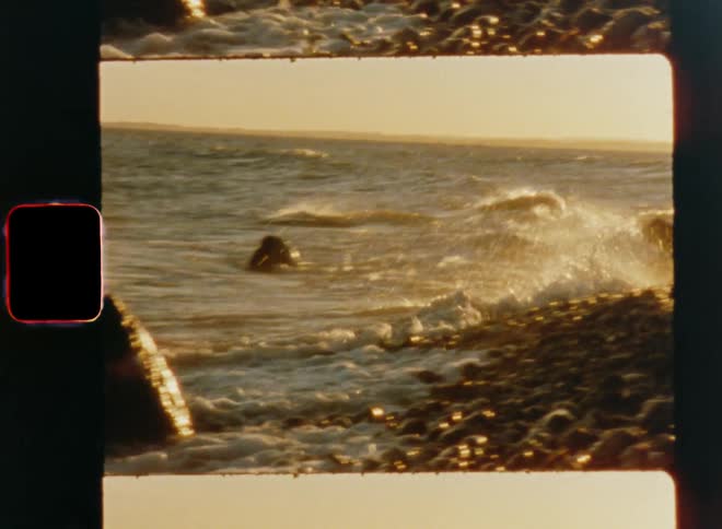 Ocean waves wash into a rock and splash as they hit the shore or gravelly beach in the evening of a sunny day with a bright sky and golden light