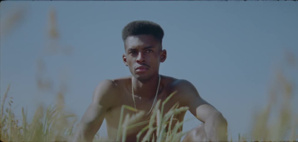 A shirtless young man sitting in a field lost in thought looks off into the distance