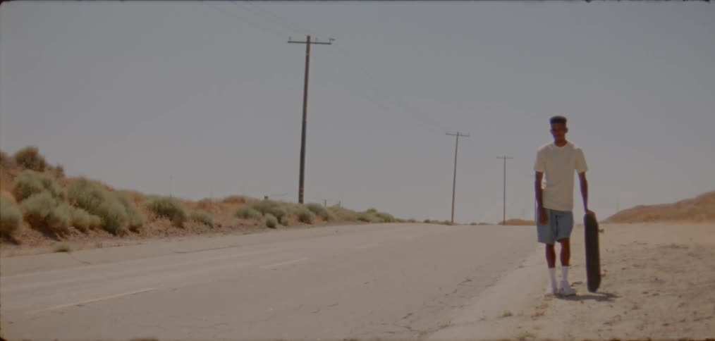 Youth stands with skateboard on side of highway in arid scrub land
