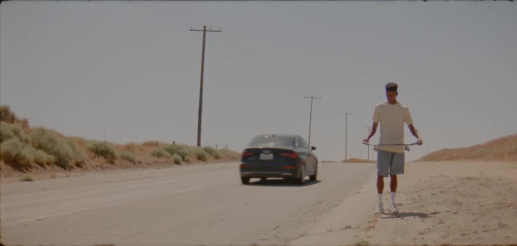 A teenager holding a skateboard stands on the side of a desert road as a car zooms past