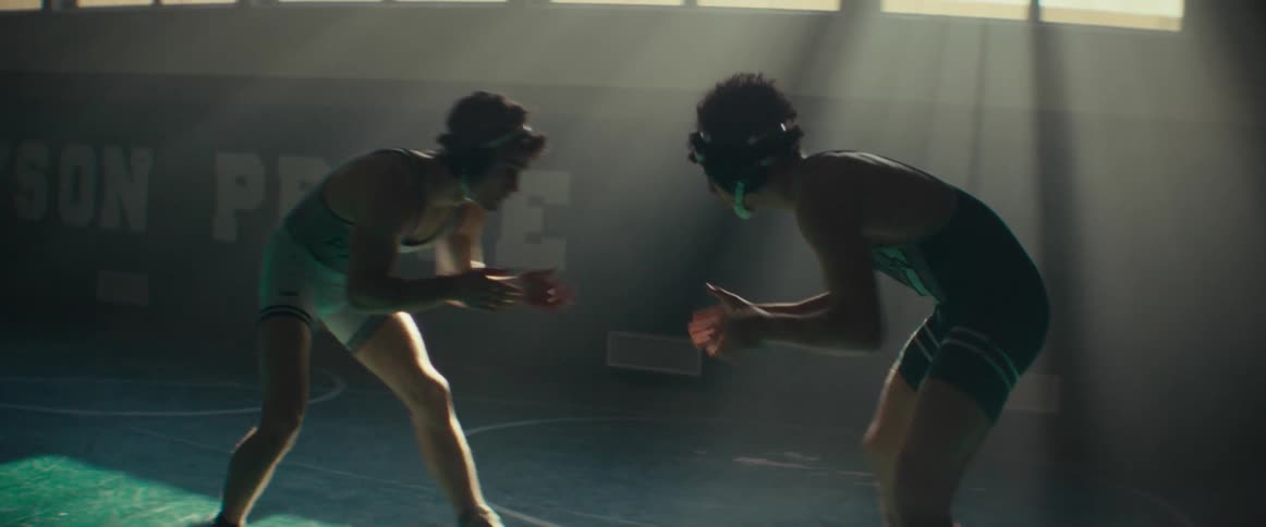 Two teenage wrestlers inside a school gym circle each other inside light coming from high windows behind them