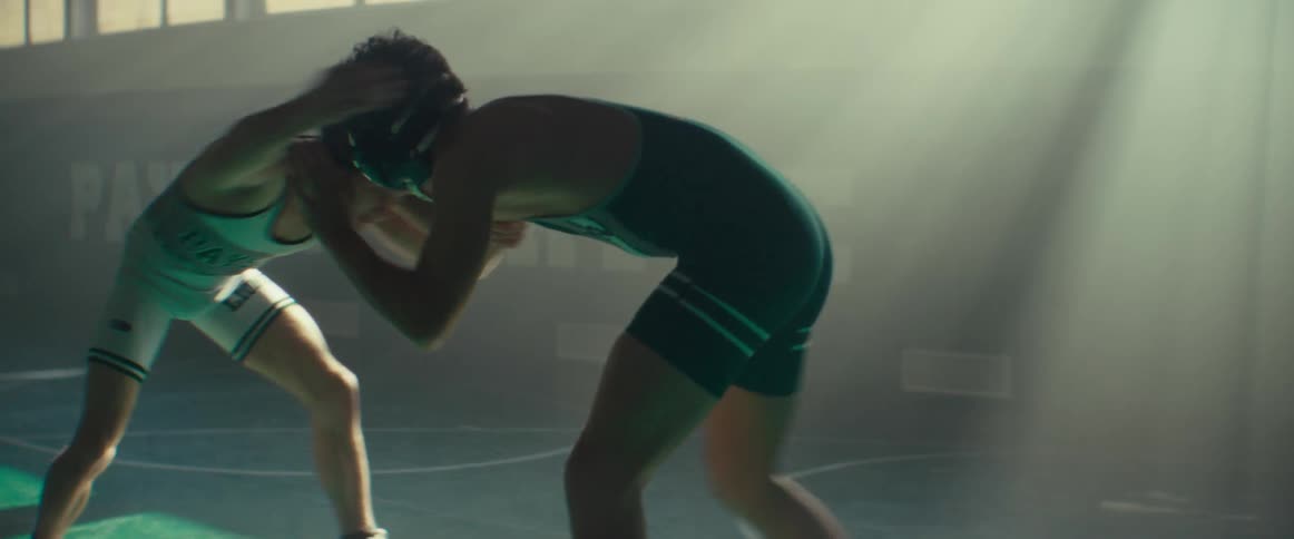 A pair of young men in wrestling singlets wrestle in a dimly lit gym during the day