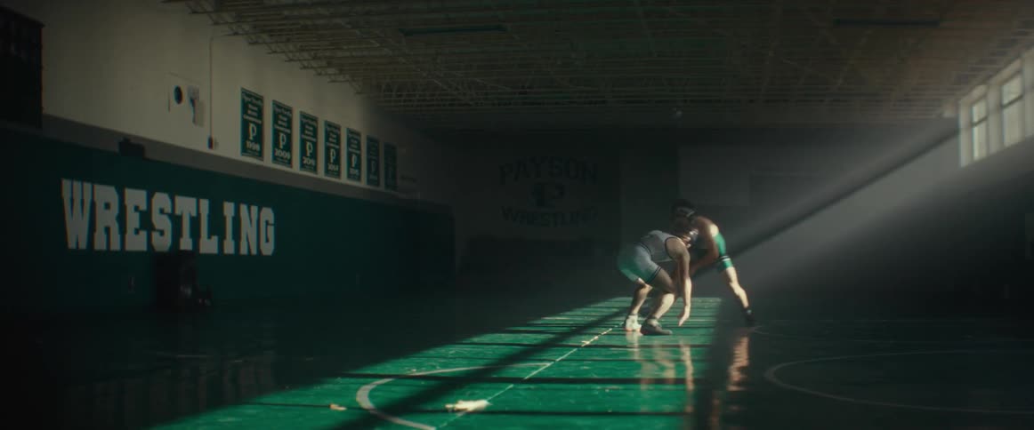 Two male athletes wrestle in dim gym lit by sunlight from high placed windows