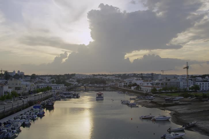 Traffic flows along road paralleling boat lined river under partly cloud covered sun