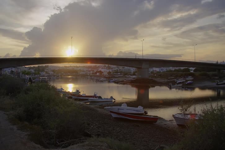 Sun shines from near horizon onto reflective river lined by boats and crossed by bridge
