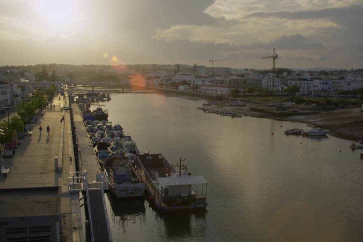 Boats of different sizes are parked by the river as few people are strolling