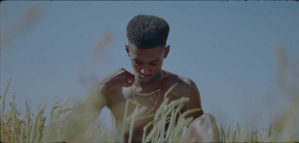 A shirtless man adjusts his necklace while sitting in a field during the day