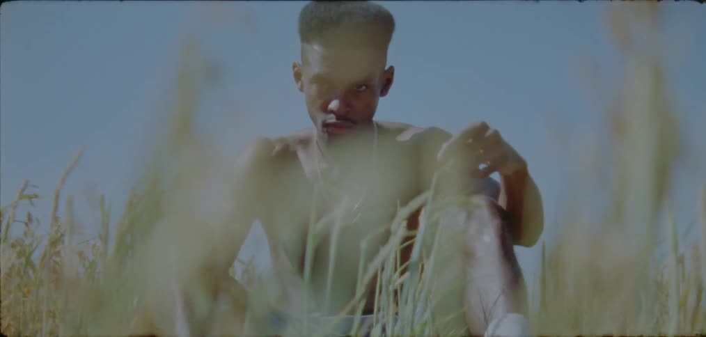 Shirtless young black man wearing a necklace sitting on the grass alone on a sunny day