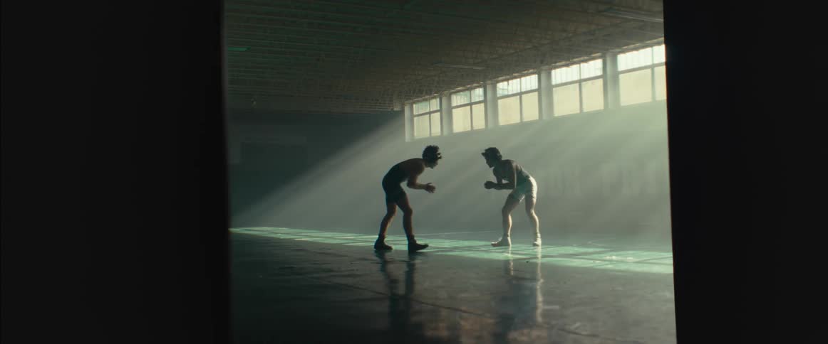 A pair of wrestlers in protective gear circle each other in a dimly lit gymnasium during the day