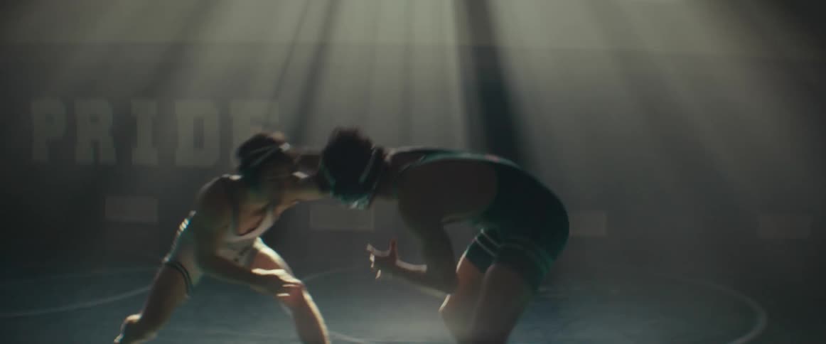 A pair of young men in singlets wrestle in a dimly lit gym during the day