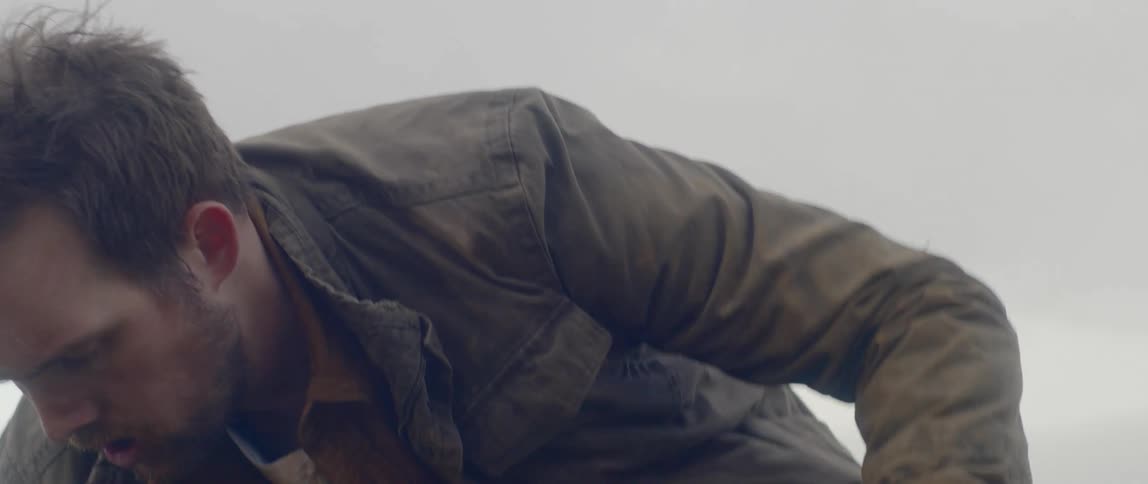 A white man squats on the ground while holding tent stakes