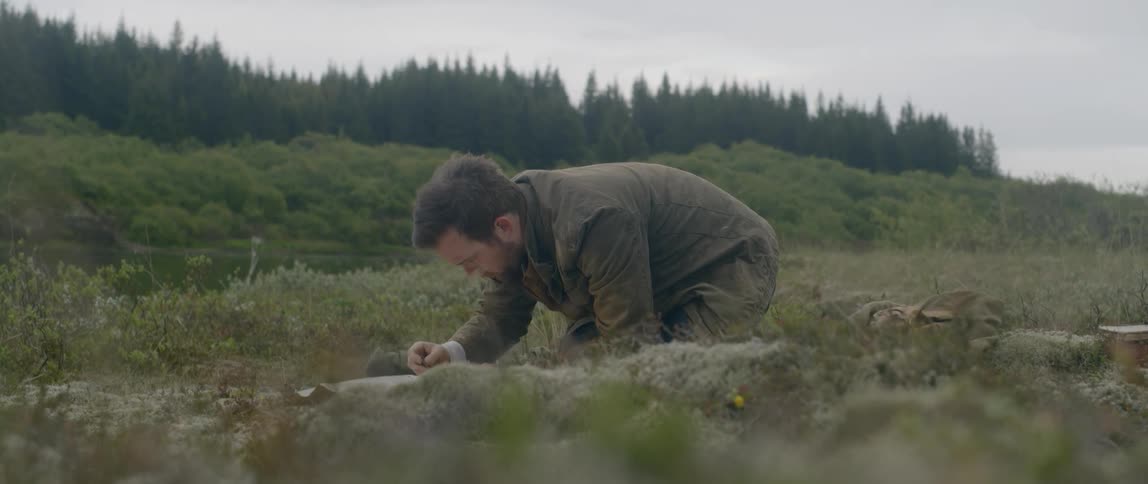 Man kneels on ground writing on paper with forest on horizon