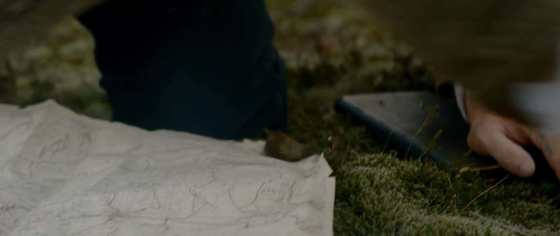 With urgency and determination a bearded man spreads out a map on the ground outside and places rocks at the corners in order to weigh it down