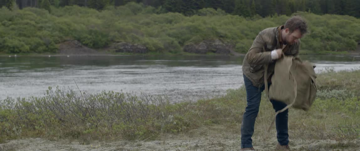 A man walking away from a river removes a backpack to place on the ground and immediately begins going through it as if hes looking for something specific