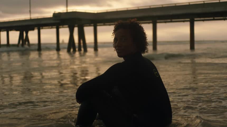 A person in a wetsuit sits in the tide on the beach while smiling into the camera on a cloudy evening