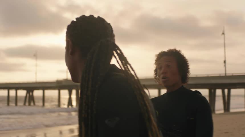 Woman teaching a woman to surf on a beach with a pier on the horizon