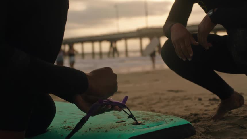 A surfer ties a leash string to a surfboard over the sand in a beach at evening