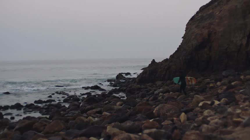 A surfer carefully walks across a beach covered in rocks towards the ocean while carrying a surfboard