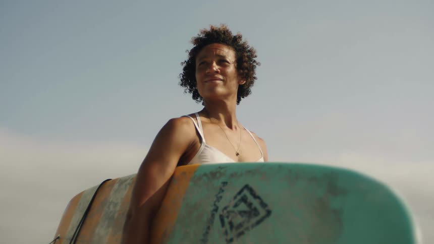 A woman holding a surfboard under one arm looks around at her surroundings while standing outdoors during the day