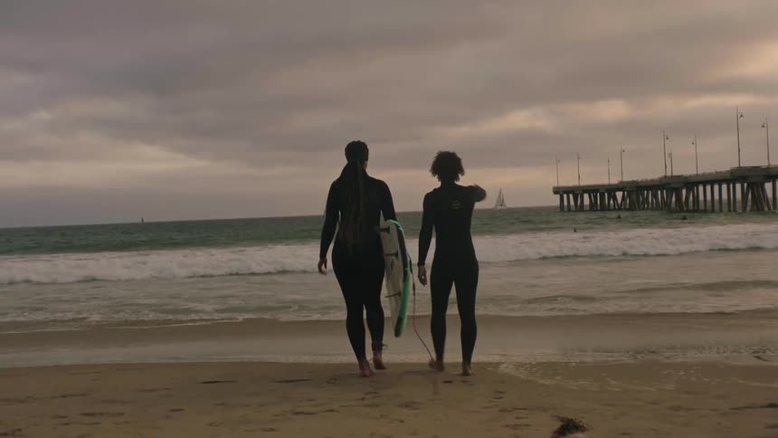 A surfer gestures at the ocean while walking with another surfer towards the water on a cloudy day