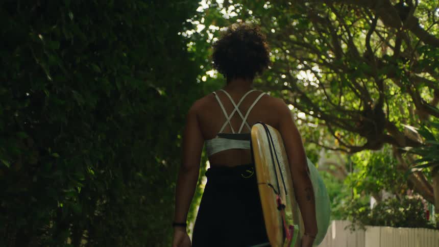 Woman walking through a treelined landscape with a surfboard in her arm