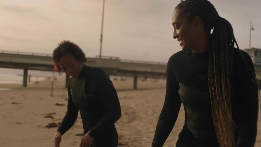 One woman instructs another in surfing while standing on beach