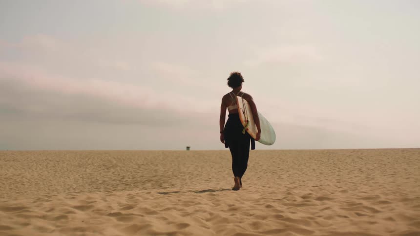 A woman holds her surfboard as she walks on the sandy beach towards the water