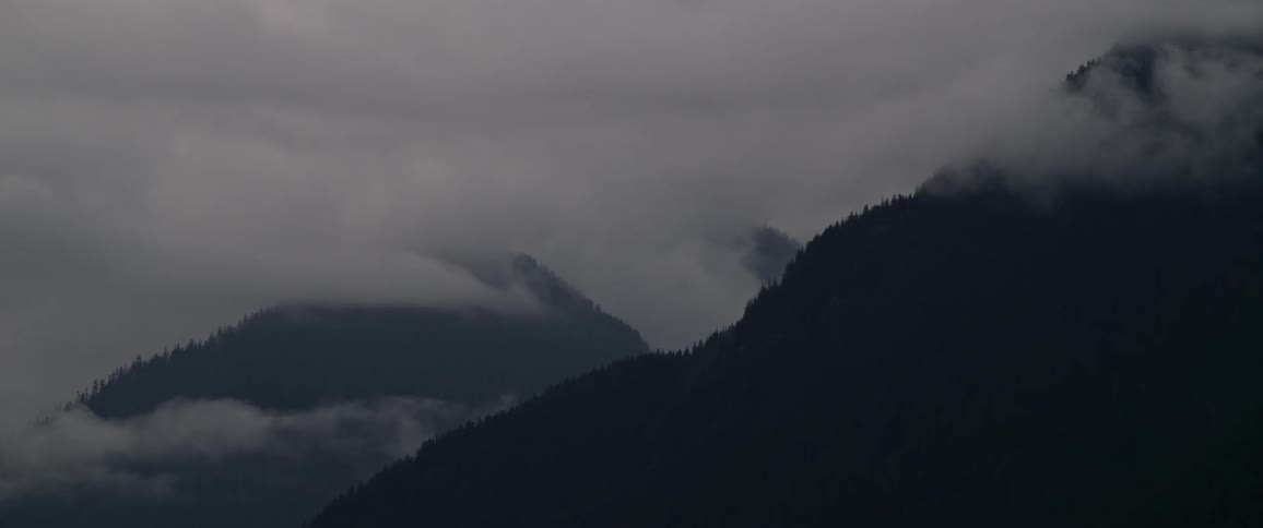 A forest covered mountain range shrouded by clouds during the day