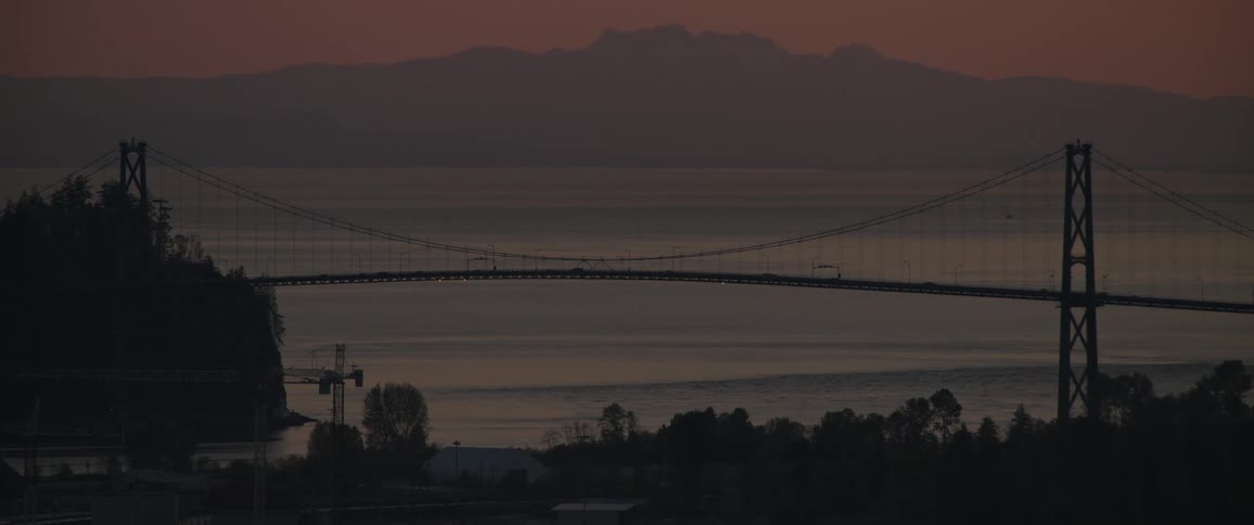 Waterfront buildings and suspension bridge are silhouetted against water on foggy day under a red sky