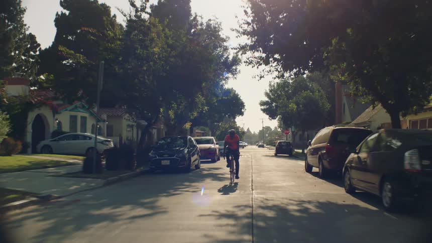 Cyclist pedals down residential street lined by cars on sunny day