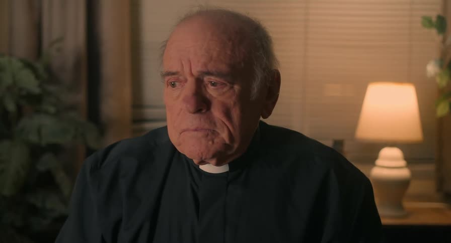 An elderly priest with a worried expression on his face makes the sign of the cross over his chest before gazing upwards