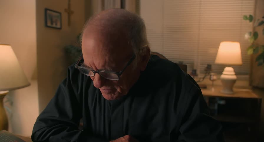 An elderly man sits at home and contemplates as he looks down at a table