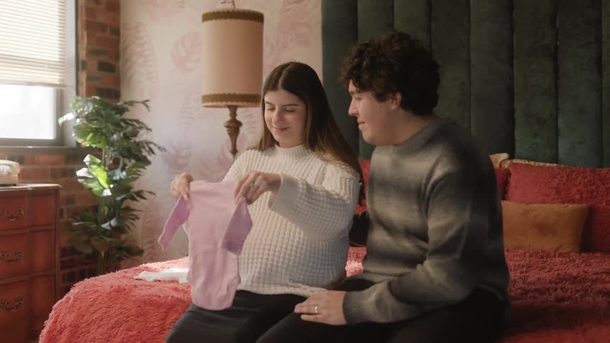 Expectant parents to be smile while sitting on bed and looking at baby clothes
