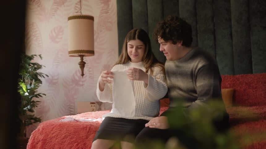 Young man and woman sitting on the edge of a bed while looking at baby clothes and smiling