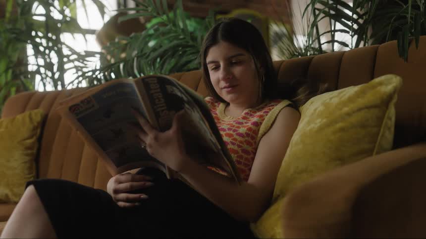 A pregnant woman strokes her belly while reading a magazine on a sofa during the day