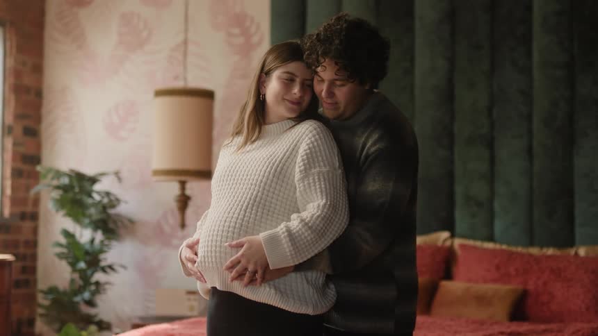 A man kisses a pregnant woman on the cheek while hugging her from behind