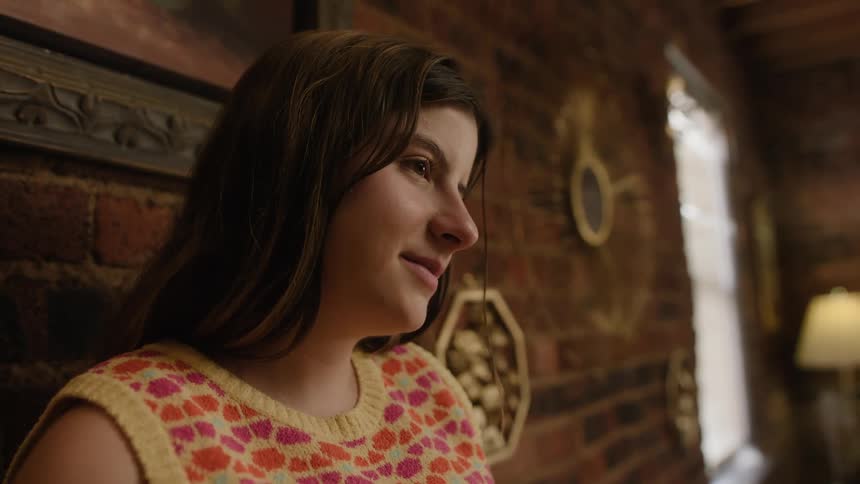 Thoughtful young white woman leaning against the wall near a window wearing a gentle smile