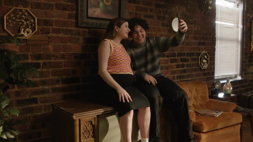 A couple sits atop a tube tv joyfully engaged in a video call on a smartphone proudly displaying the womans pregnant belly