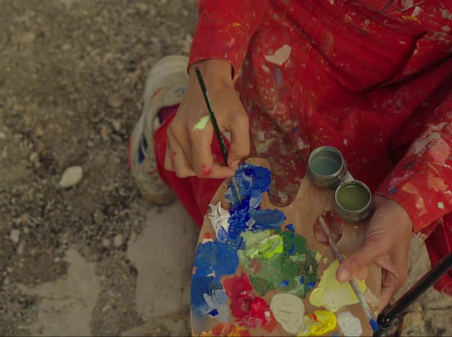 A woman uses a paint brush to pick blue paint off a messy palette