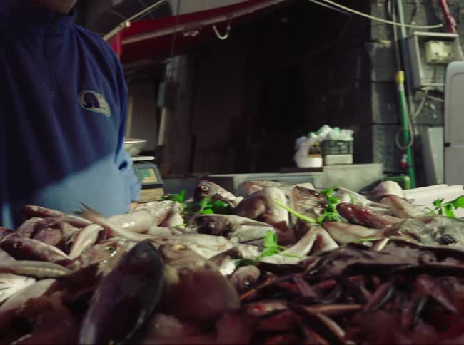A man pointing to fish on a table and in the back ground a man carrying boxes