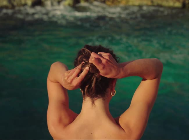 A woman standing in water puts her hair in a bun puts the top of her bathing suit back on and walks away in the water